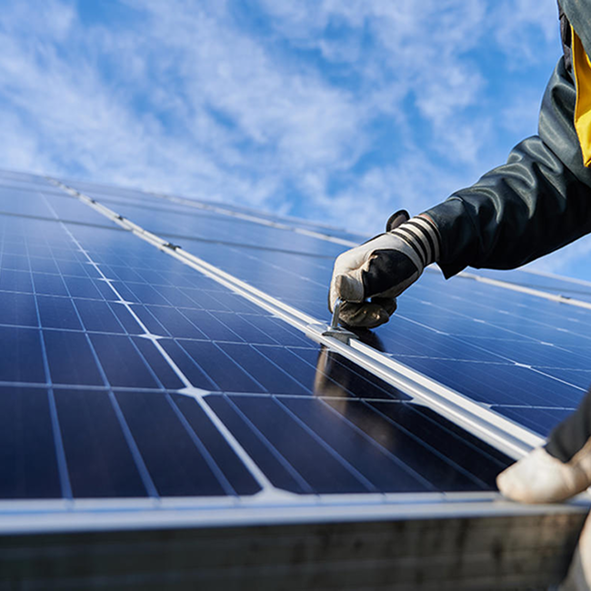 Mano protetta da un guanto lavora su un pannello fotovoltaico, sullo sfondo cielo sereno con nuvole sparse 