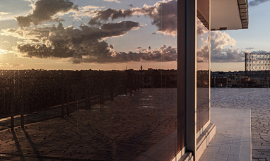 Dettaglio della terrazza della sede del Gruppo Acea al tramonto, con il sole riflesso sulla vetrata sulla sinistra, e con vista sulla città di Roma e il Gazometro sullo sfondo.