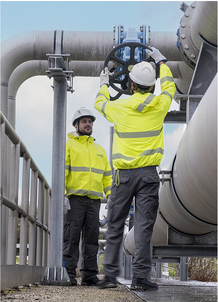 Due operai con caschi e giacche ad alta visibilità lavorano a una grossa tubatura industriale all'aperto