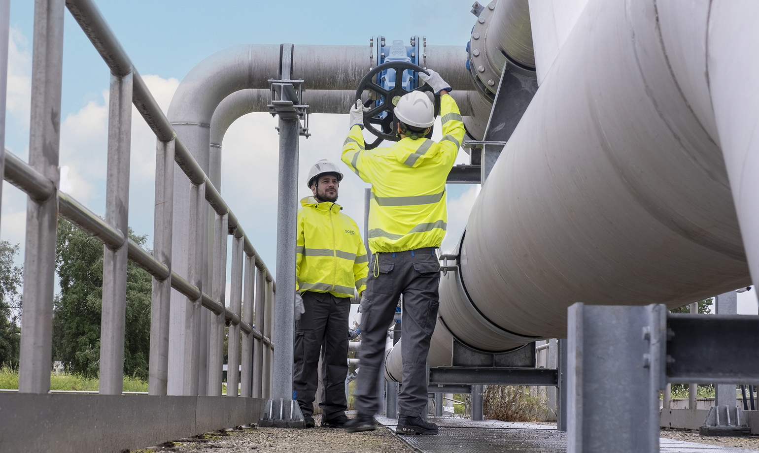 Due operai con caschi e giacche ad alta visibilità lavorano a una grossa tubatura industriale all'aperto