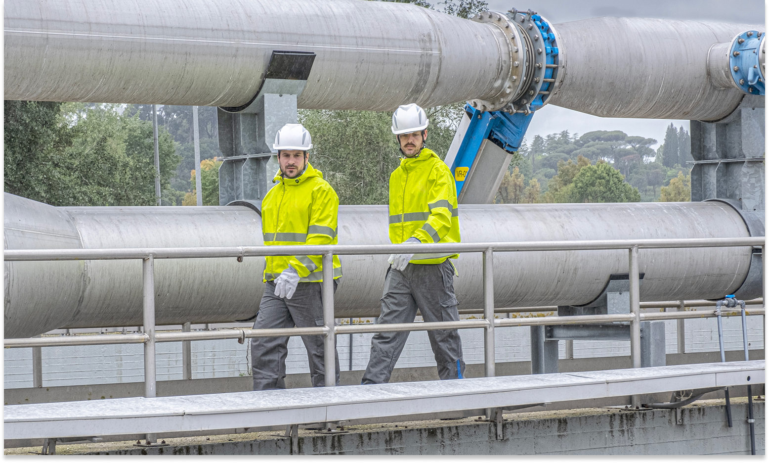 Due operai con caschi e giacche ad alta visibilità camminano su una passerella in un impianto industriale con grandi tubature metalliche sullo sfondo