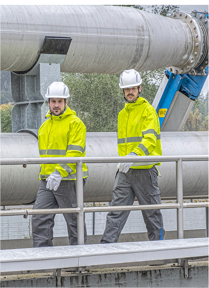 Due operai con caschi e giacche ad alta visibilità camminano su una passerella in un impianto industriale con grandi tubature metalliche sullo sfondo
