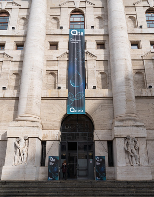 Un grande edificio reca un banner verticale con il logo di Acea e il testo "28" in alto, seguito da "Piano Industriale 2024-2028" e un logo con "Acea" in basso.