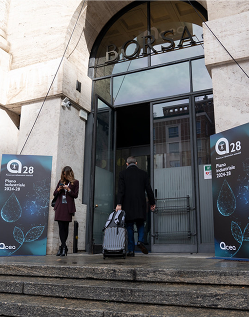 Due persone davanti a un edificio: una guarda il telefono. Ai lati dell'ingresso ci sono due banner con il testo "Acea 28 Piano Industriale 2024-28".