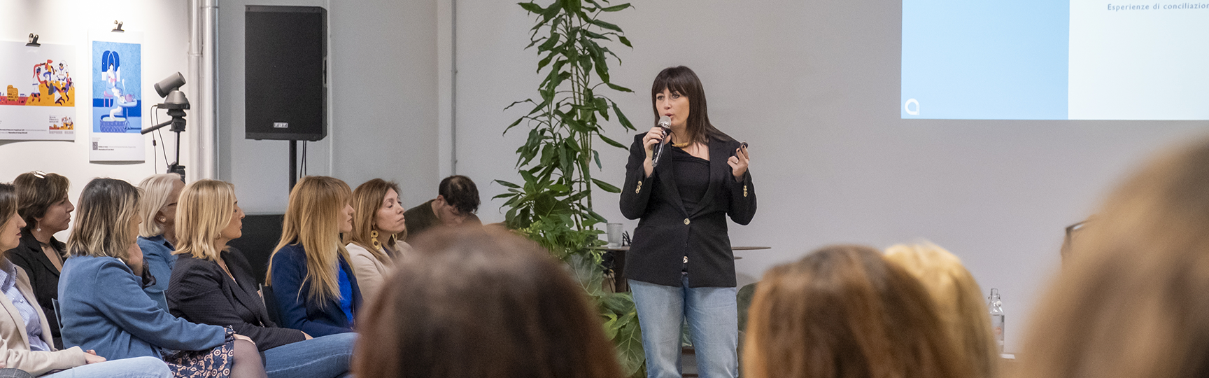Una classe di persone assiste ad una lezione. Si intravede una slide proiettata e c'è un docente che parla tenendo in mano uno strumento di lavoro, oggetto della lezione