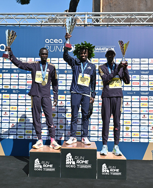 Atleti alla partenza della ACEA Run Rome the Marathon 2024 sotto l'arco di arrivo con coriandoli tricolore che cadono, atmosfera festosa con cielo azzurro e monumenti di Roma sullo sfondo