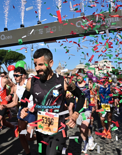 Atleti alla partenza della ACEA Run Rome the Marathon 2024 sotto l'arco con coriandoli tricolore e fontane pirotecniche, atmosfera festosa con cielo azzurro