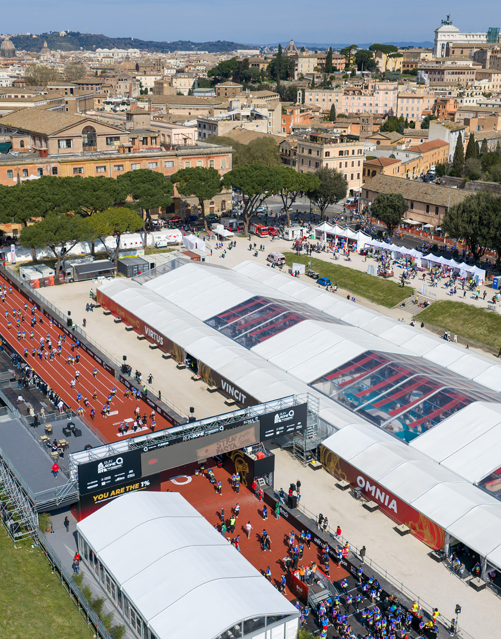 Aerial view of the Acea Run Rome The Marathon 2026 village at the Circus Maximus, featuring the red finish line, the white sponsor pavilions, and the Rome skyline in the background.