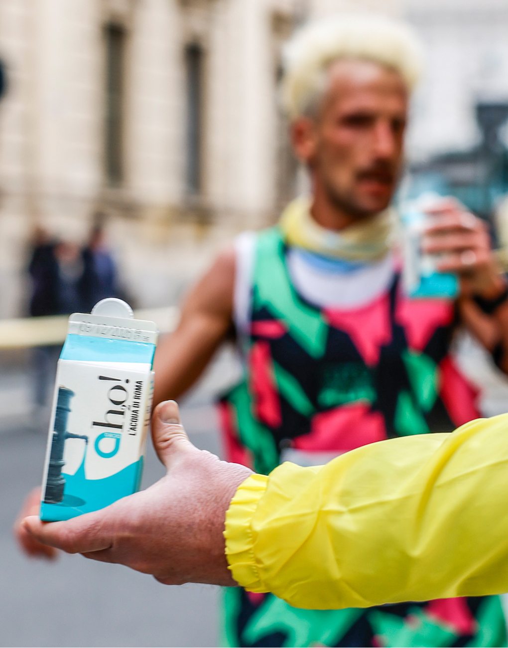 A hand is holding a carton of Ah2o! water. In the background, a runner is drinking from another carton.