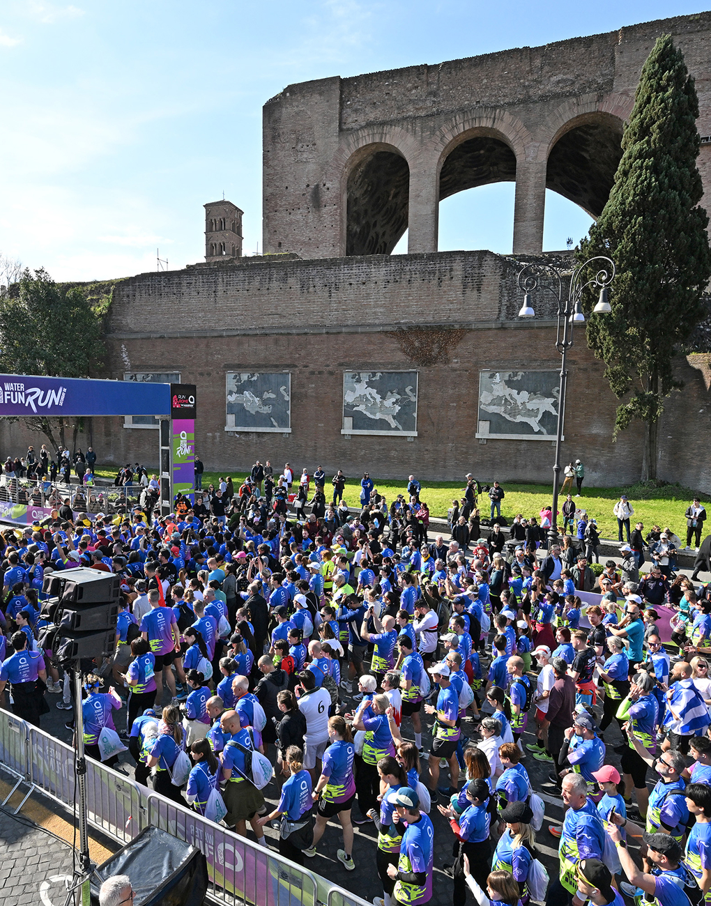 Hundreds of participants in blue T-shirts gather under the starting arch of the Water Fun Run, a non-competitive race as part of the Acea Run Rome The Marathon 2026, with the Baths of Caracalla in the background.