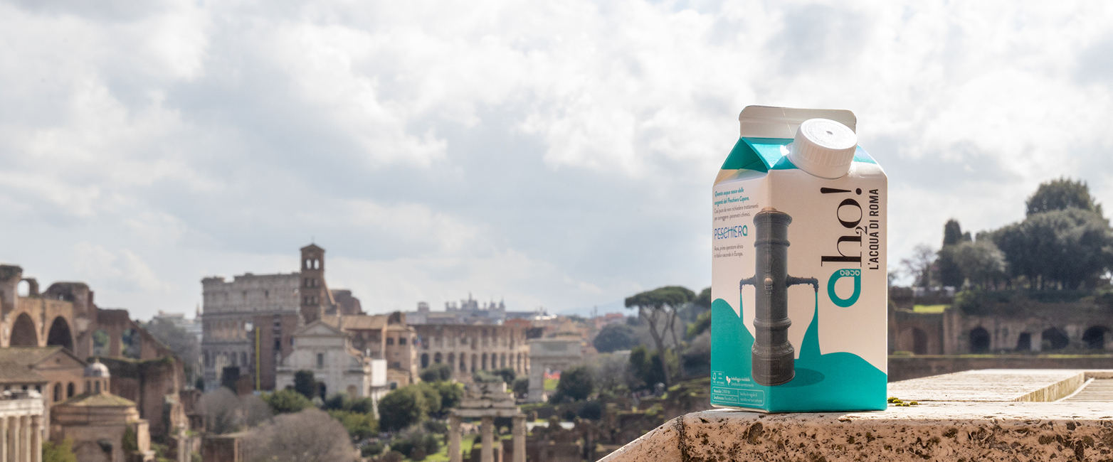 una confezione di "aho! L'acqua di Roma" di Acea, in primo piano su un muretto con il panorama del Foro Romano sullo sfondo.