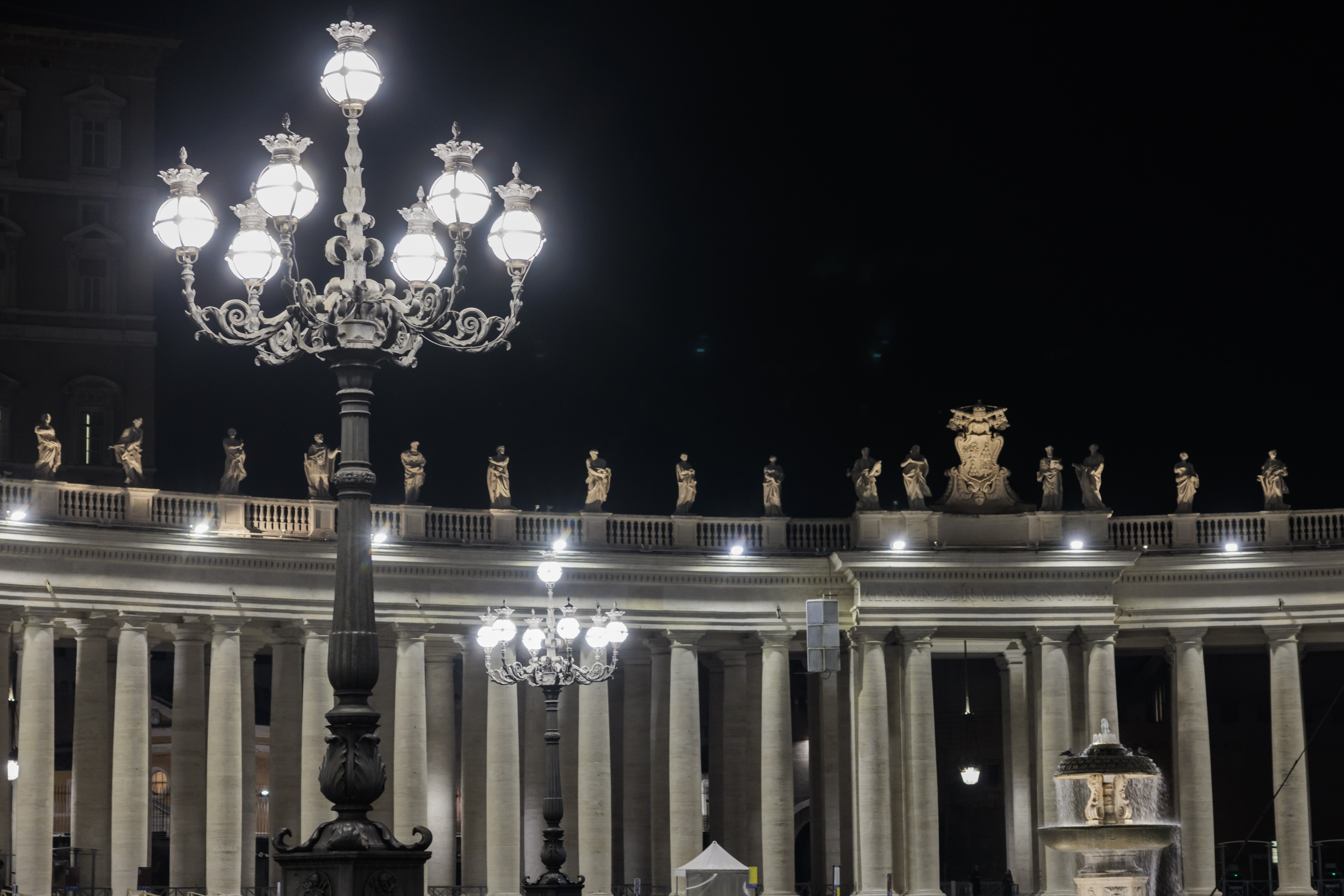 Un lampione acceso in primo piano; in secondo piano si vede il colonnato di Piazza San Pietro illuminato