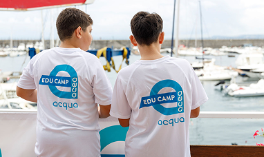 Due bambini osservano delle piccole imbarcazioni con le spalle rivolte verso l'obiettivo indossano una tshirt con il logo Acea Acqua Edu Camp